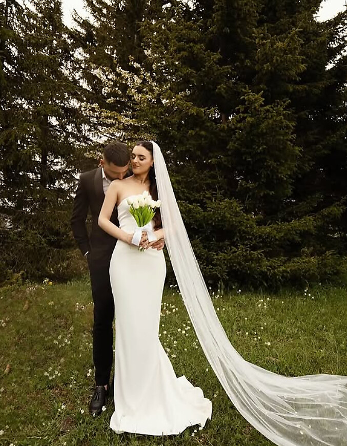 Bride in a fitted strapless gown with a cathedral veil, standing with her partner outdoors